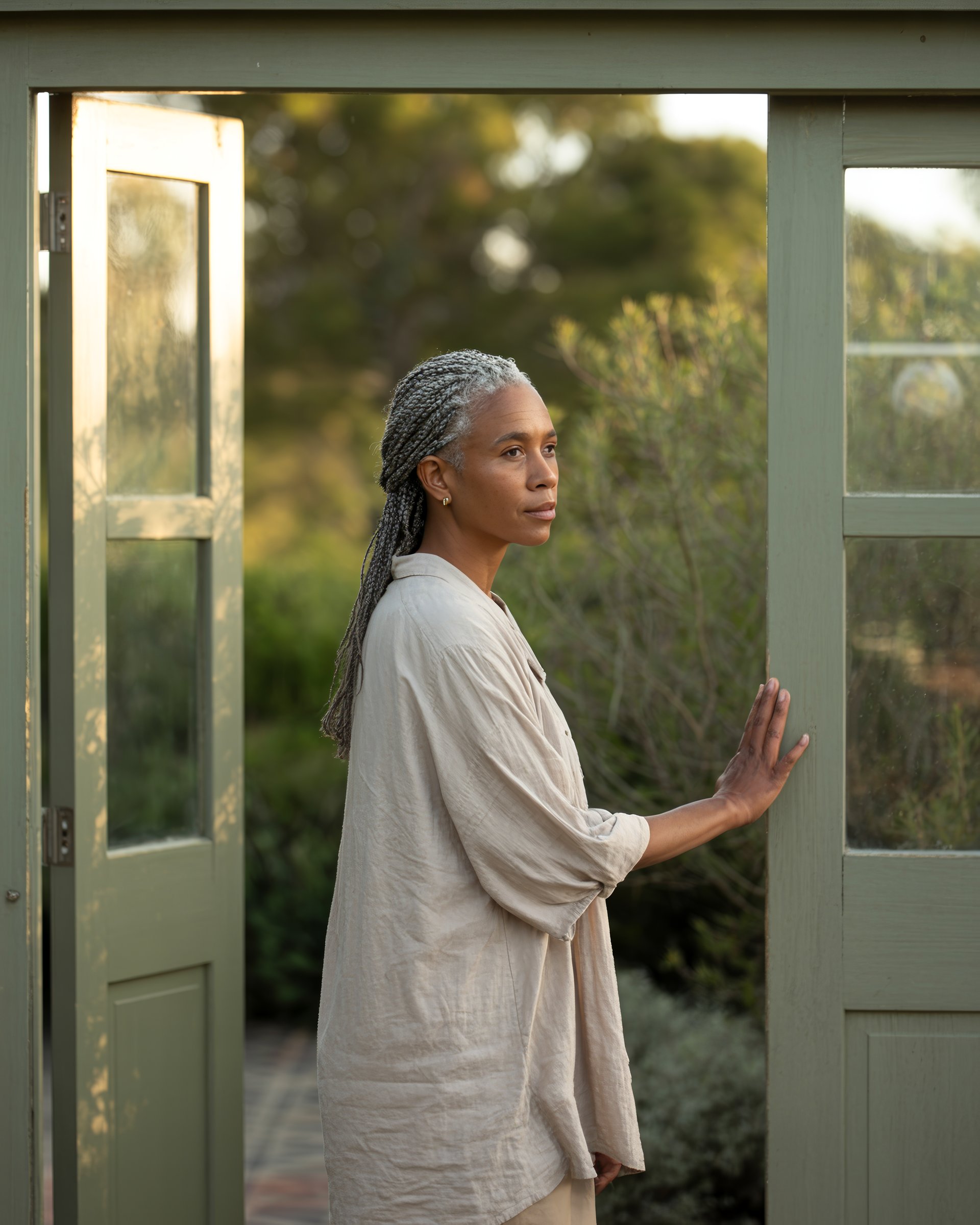Miriam Sobukwe, a Black South African woman with silver braided hair in cream linen, standing in the open frame of a sage-green Cape Dutch door